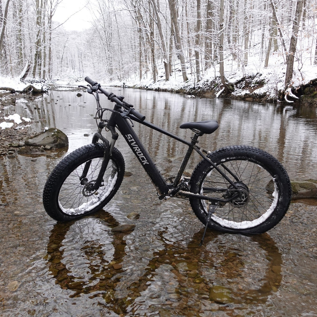 I Got It First 1000W electric fat tire bike in black standing on a rocky shallow stream in a snowy forest